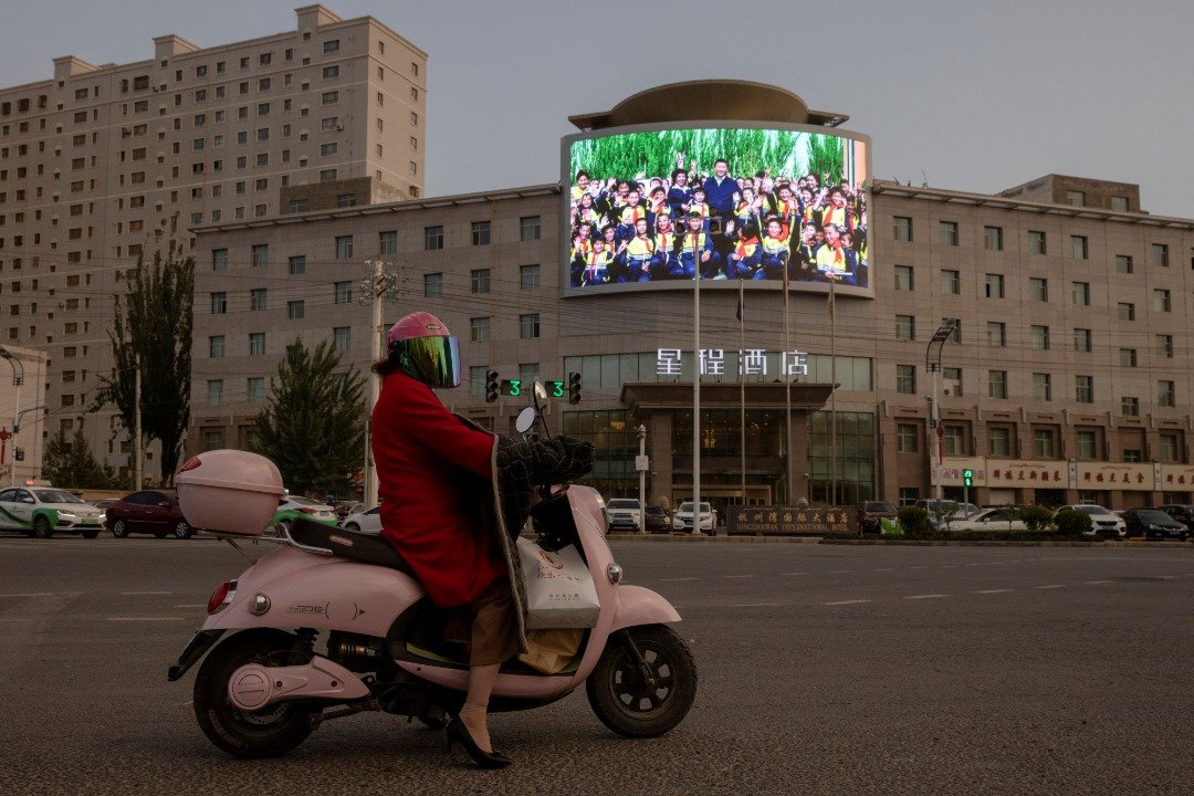 图为今年4月30日，新疆西南部和田市，一名女士骑著摩哆经过一个交通路口，酒店上的荧幕显示中国国家主席习近平与一群新疆小孩相聚一起的欢乐画面。（图取自路透社）