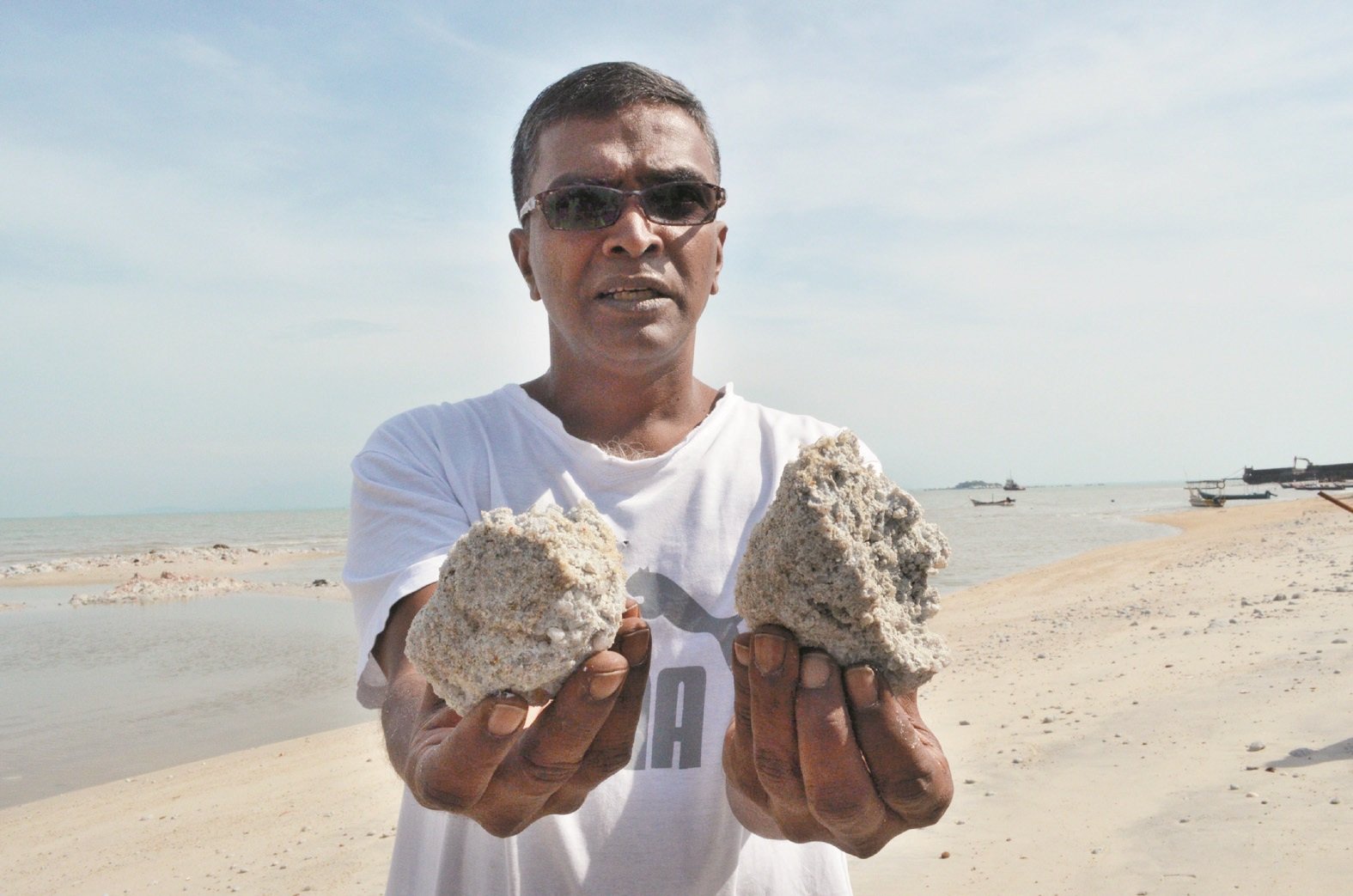 居民莫哈末丹尼尔出示结块后硬如石头的淤泥，洒落满沙滩，造成渔船危险。