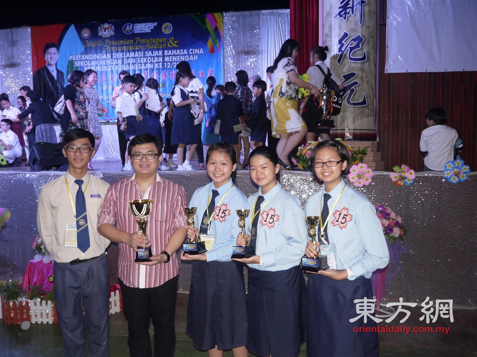 2015年第十二届全国中小学华语诗歌朗诵比赛，通过手机短讯向何书卫学习的育华中学代表（左）获得高中组季军，吉隆坡中华中学代表则获得初中组冠军。他们的奖杯包含了何书卫的血汗。