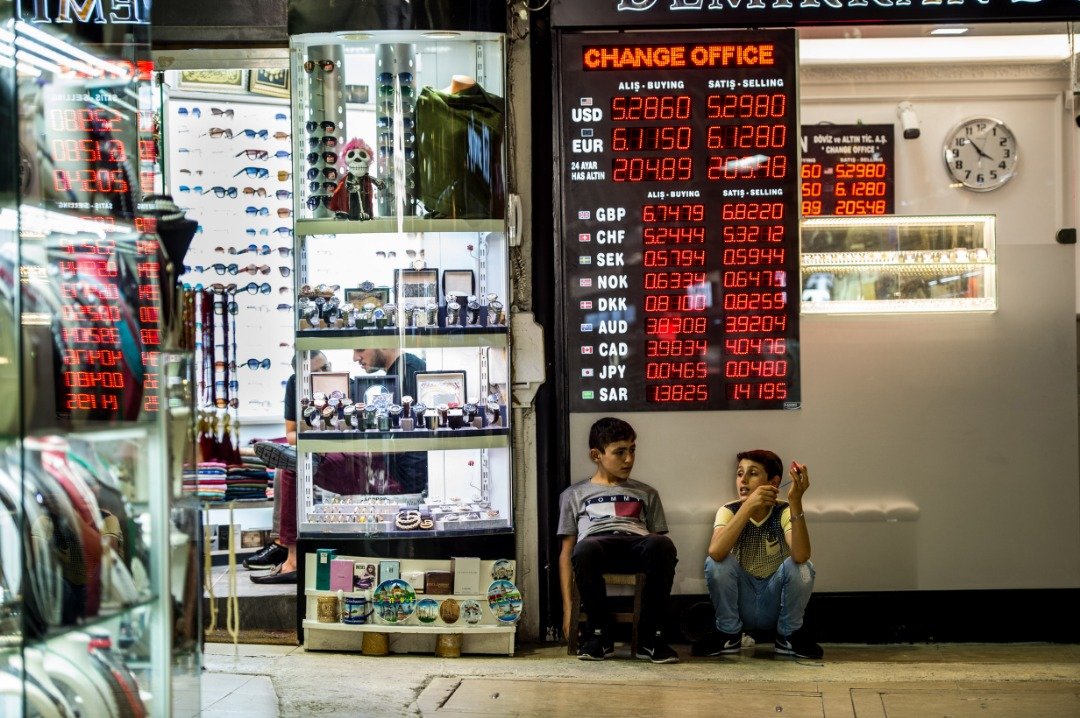 今年以来，土耳其里拉经历数次快速贬值。周三，两名男孩蹲坐在伊斯坦布尔市一家货币兑换店外，电子萤幕显示，当天5.29里拉可兑换1美元。