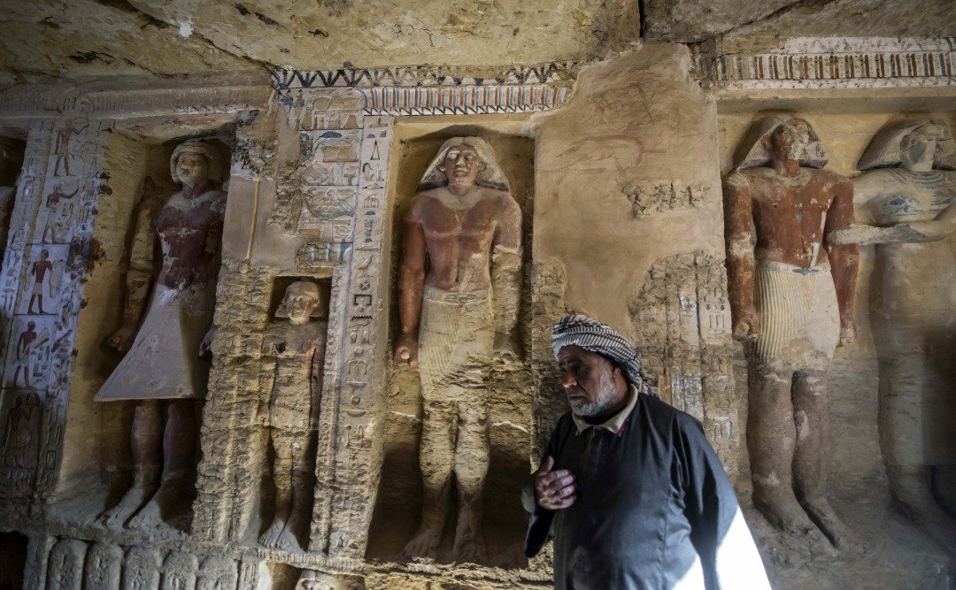 埃及考古工作人员塞阿布杜阿，站在墓葬内部，一旁的墙上刻有一些人像雕像，包括瓦提耶本人、其母亲、妻子以及其他亲属。