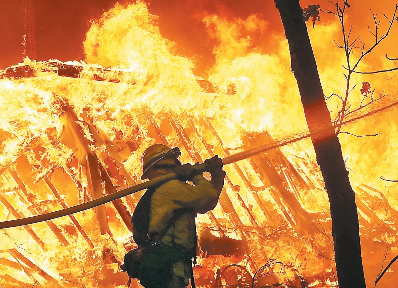 到灾场救火的加州消防人员，扛著消防喉往大火喷洒。其身旁的房 子被烈火吞咽。