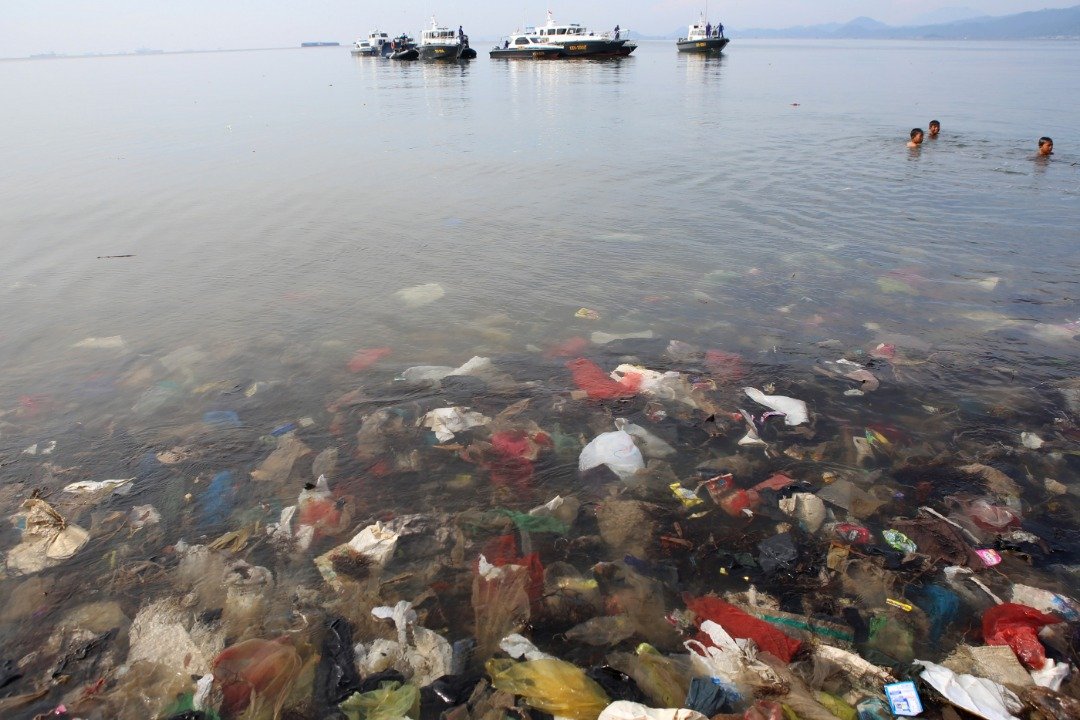 为了减少囤积在海边的海洋垃圾，印尼政府策划了各种环保措施。这是淹埋在班达楠榜海边的海洋垃圾，一旁还有孩童在水中游泳玩乐，不远处亦有几艘军人的船只。