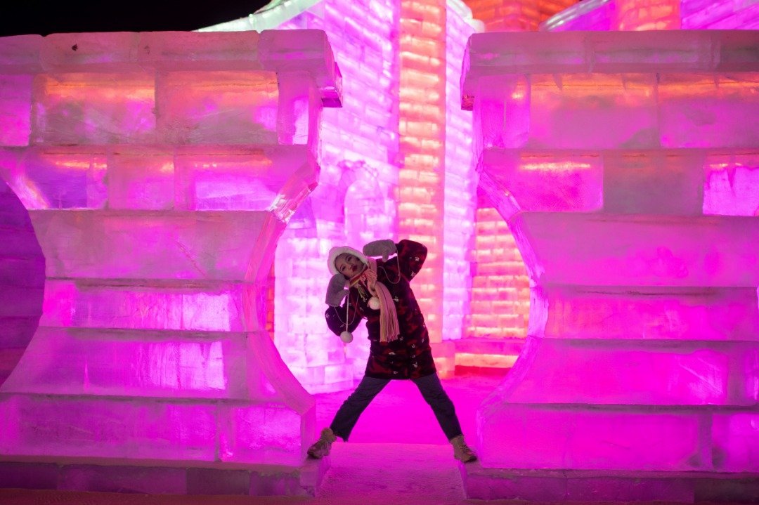 一名游客在被粉紫灯光映照得如梦似幻的冰雕前，摆著姿势拍照留念。