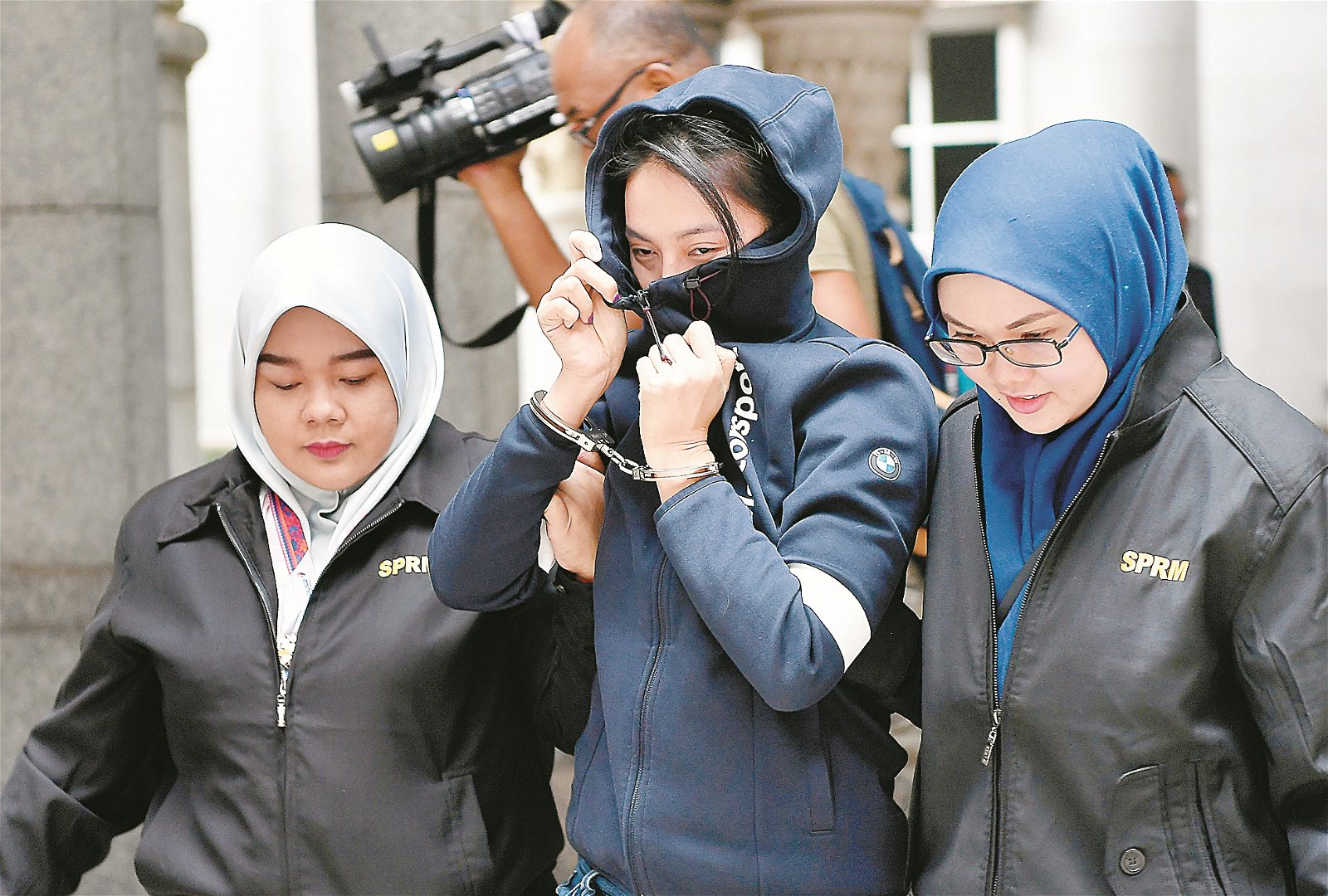 私人公司女首席执行员简芝颖（中），反贪会官员的护送下带入推事庭时，用衣服闪避摄影的镜头，而其双手被套上手铐。