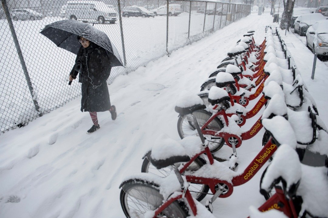 大雪中，一名妇女周日撑著伞走在华盛顿的街头，路旁并排的共享脚车上堆满了厚厚的一层积雪。