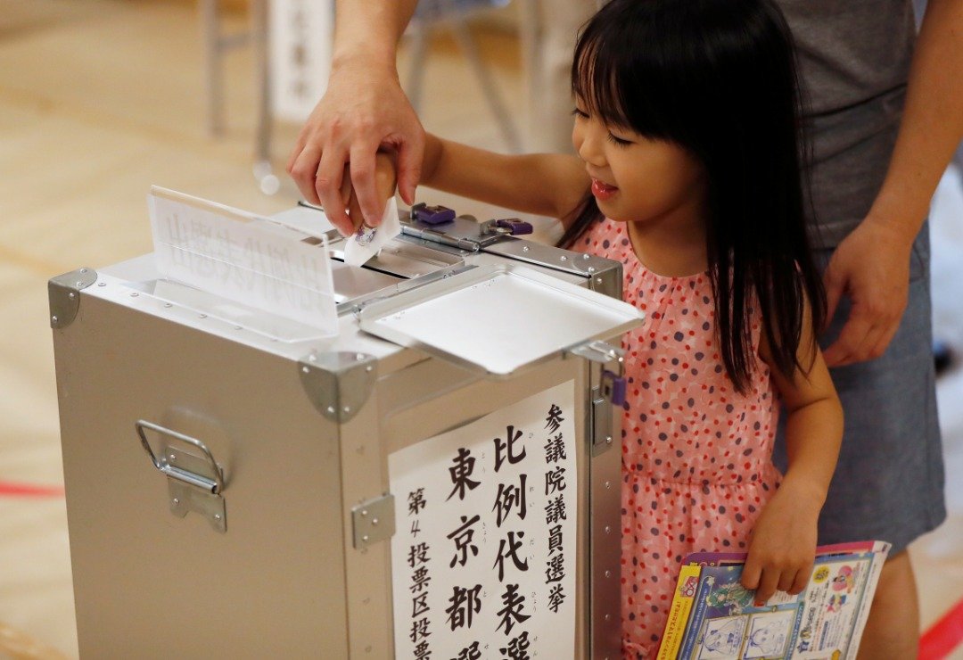 东京市民周日大早前往投票站投下神圣一票，一名小女孩贪玩地协助家长将选票投入票箱中。