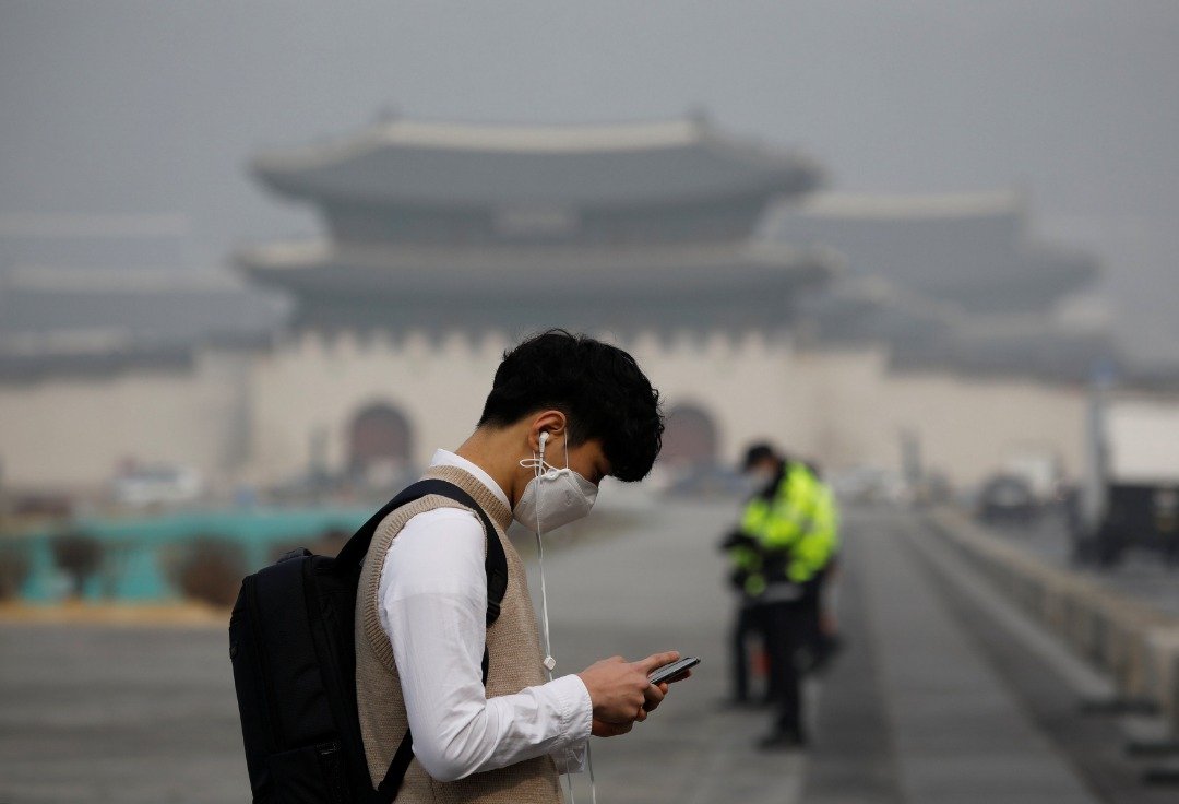 一名学生戴著口罩，低著头看手机，走过光化门广场。不远处的地标景福宫笼罩在雾霾中，一片朦胧。