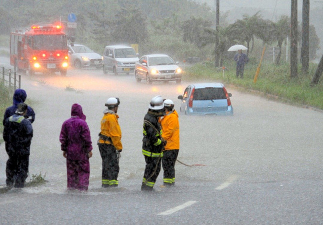 洪水淹没了道路低洼区，导致交通工具无法继续前行，一辆轿车更是被洪水包围。而受困在一旁的民众，正等待前来的消防人员施予救援。