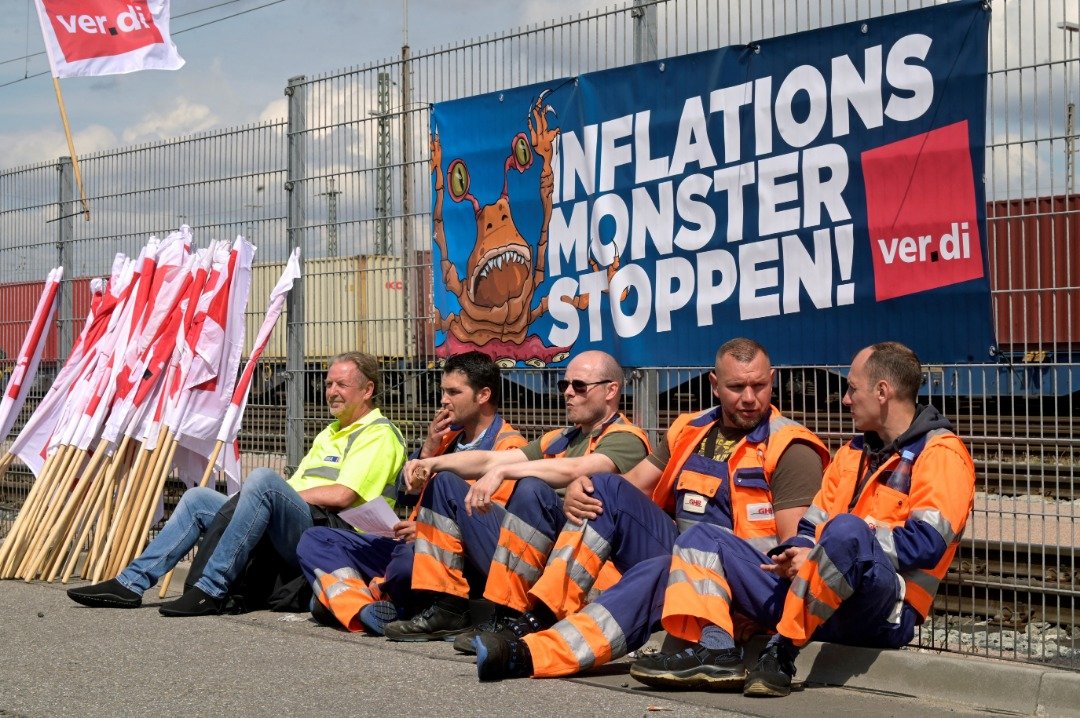 今年6月9日在德国汉堡港举行罢工以争取更高工资时，工人们坐在Burchardkai集装箱码头的横幅前，上面写著“阻止通货膨胀怪物”。（图取自路透社档案照）