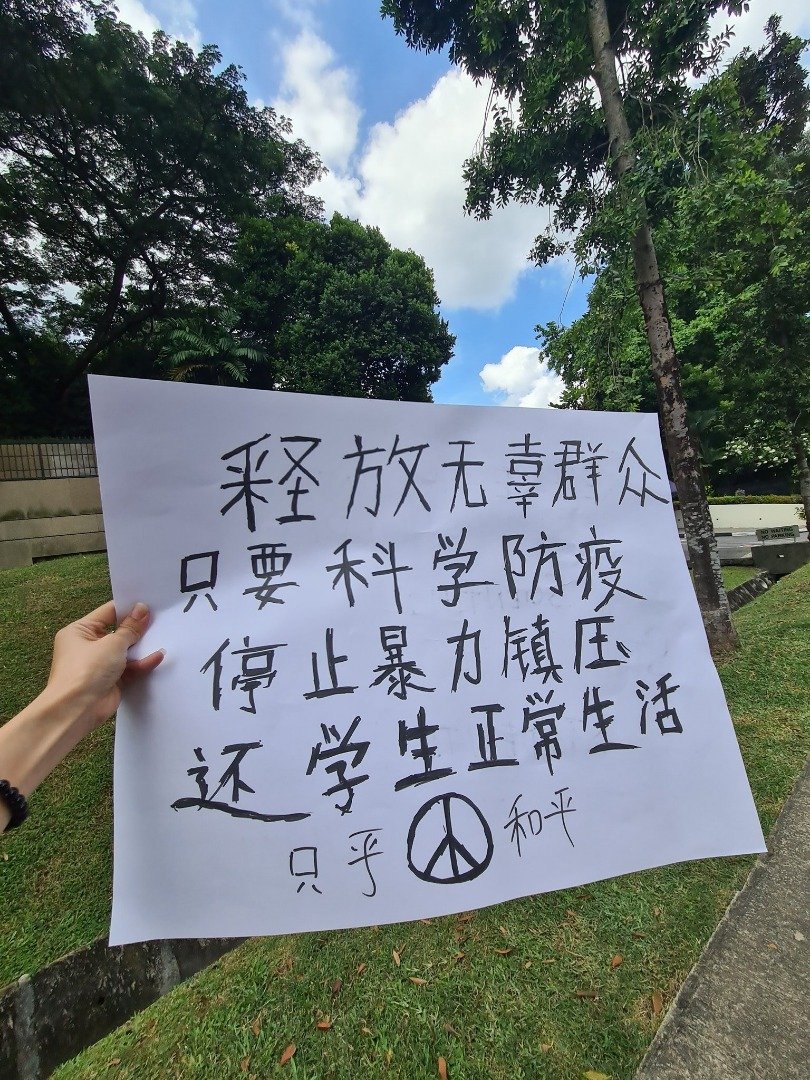 涉案女子在中国驻新加坡大使馆前举抗议标语，写着“释放无辜群众、只要科学防疫；停止暴力镇压，还学生正常生活。”