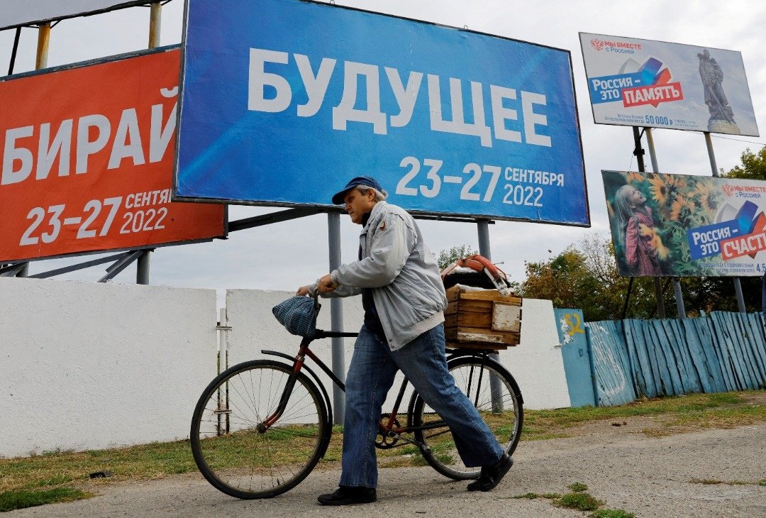 周一，在扎波罗热州，由俄军控制的梅利托波尔市，男子推著自行车经过写有“未来 2022年9月23日至27日”的投票横幅底下。（图取自路透社）