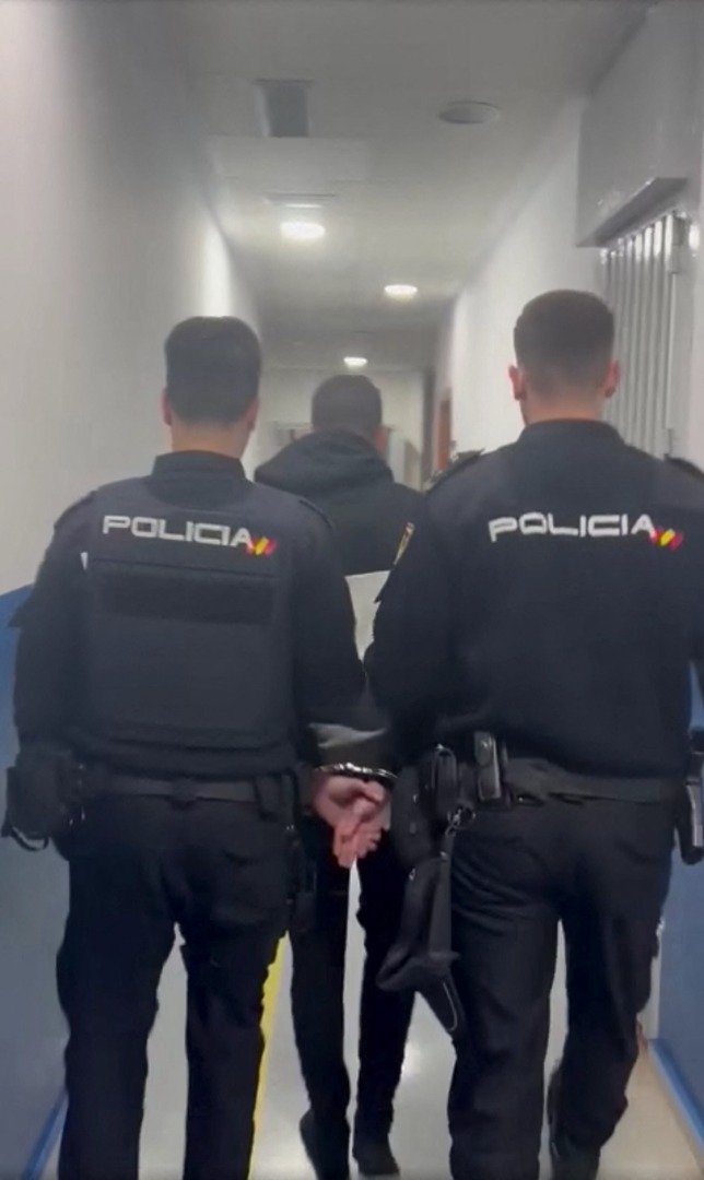 西班牙警方公布的一段视频画面显示，两名警察护送一名身著连帽运动上衣、戴著手铐的男子到警局。（图取自西班牙警方/路透社）