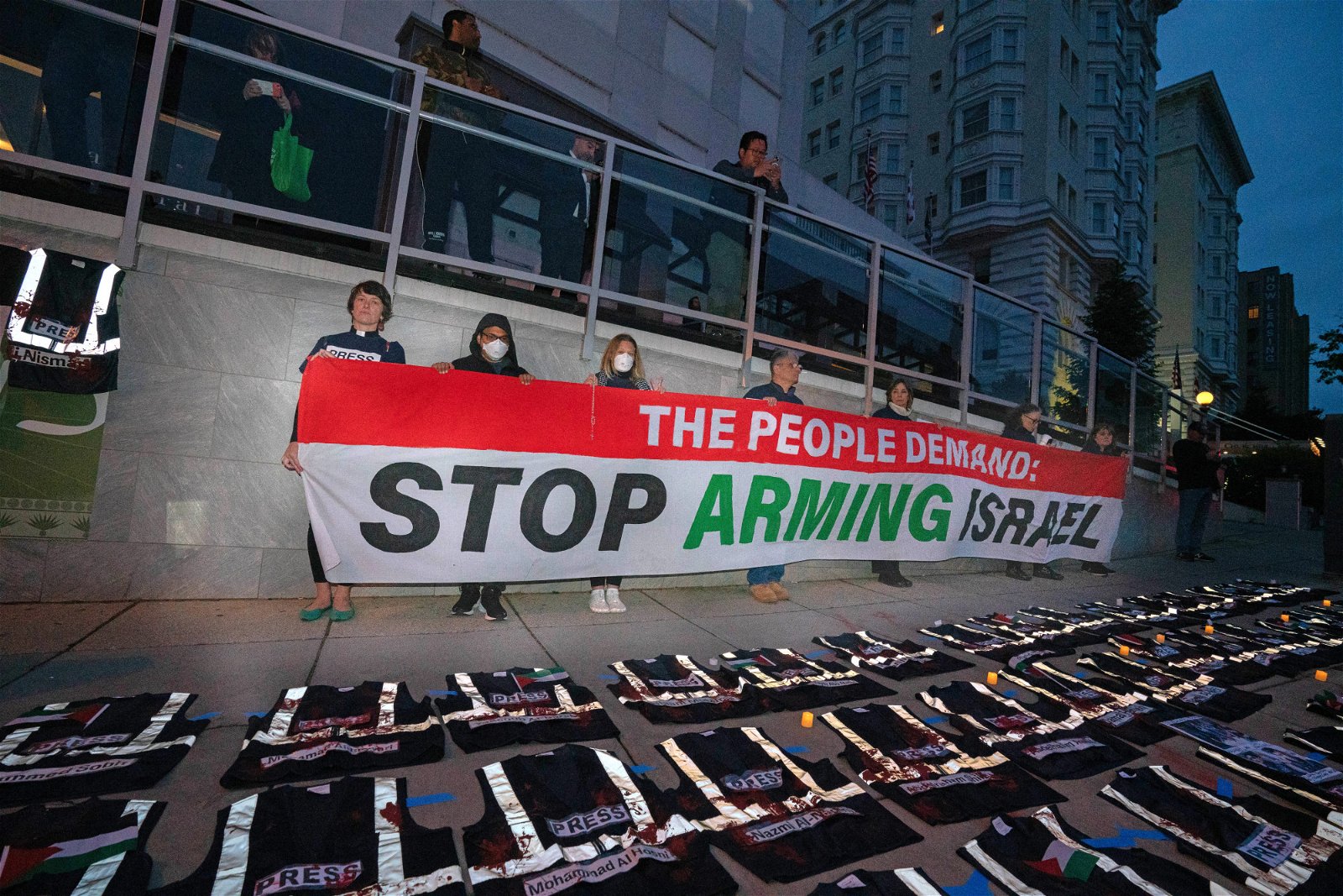 周六在华盛顿希尔顿酒店外，抗议者手持“停止武装以色列”标语横幅示威，并在人行道上为被杀害巴勒斯坦记者设立了临时悼念处，写有死去记者名字的防弹背心一件件铺在地上。（图取自法新社）
