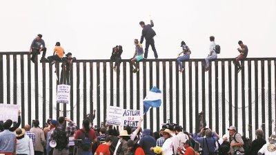 许多年轻的移民，无视执法人员警告，攀爬上边境围篱，并望向美国国境的一方。