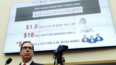 美国财长姆努钦周四到众议院金融服务委员会作证时，竭力缓和议员们对特朗普贸易政策的担忧。其身后的大萤幕显示美国目前债务的数额。