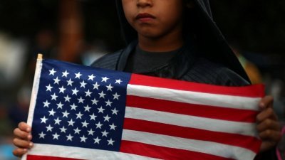 来自洪都拉斯的8岁男童米兰达，在埃尔查帕拉尔的美墨入境点外，拿著美国国旗面对墨西哥警方，希望当局放行让他和家人进入美国。