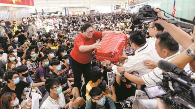 一名身穿红衣的妇女站在坐满示威者的离境大堂中，试图将手中的行李箱交给机场工作人员。受反修例示威集会影响，香港机场周二连续第2天出现瘫痪，更被迫暂停航班登机作业。