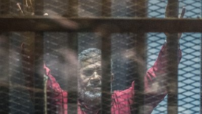 遭军方推翻的穆尔西被捕后遭囚禁多年，难以获得医疗照护。图为穆尔西2016年出庭时，在金属网围起的厚厚玻璃隔间举手向外界示意。