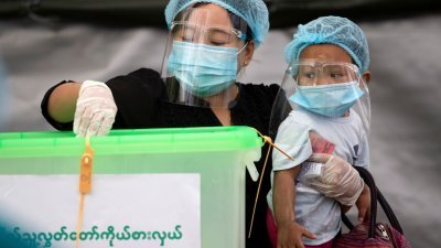 缅甸周日举行全国议会选举，当地选民一早戴著口罩前往投票站投票。为防新冠病毒传染，一名戴著口罩及面罩，做足防疫措施的妇女抱著孩子，在投票站把填好的选票投入票箱，履行公民责任。（路透社）