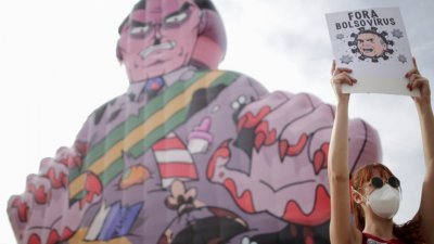 在巴西首都巴西利亚，示威者手持“博索病毒滚开”标语牌，在国会大楼外要求议员对总统博索纳罗进行弹劾，示威者背后是大型讽刺博索纳罗的塑胶人偶。（图取自路透社）