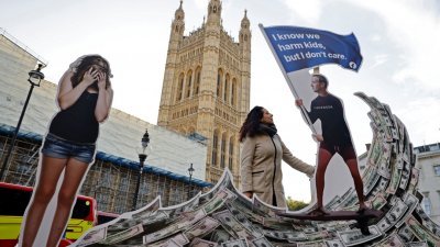 在英国伦敦市中心国会大厦对面的抗议活动中，一名示威者周一摆出一幅装置，描绘面子书创始人朱克伯格拿著一大笔现金冲浪，周围都是陷入困境的青少年。（法新社）