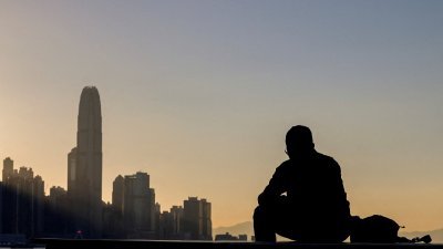 图为一名男子坐在香港维多利亚港口，背景为中央金融区。（图取自路透社）