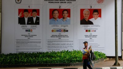 印尼在2月14日举行总统选举投票，选民们将决定谁将执掌这个东南亚最大的国家。在首都雅加达，一名携带随身物品的街头小贩走过一面横幅，横幅上显示3组正副总统候选人。（图取自路透社）